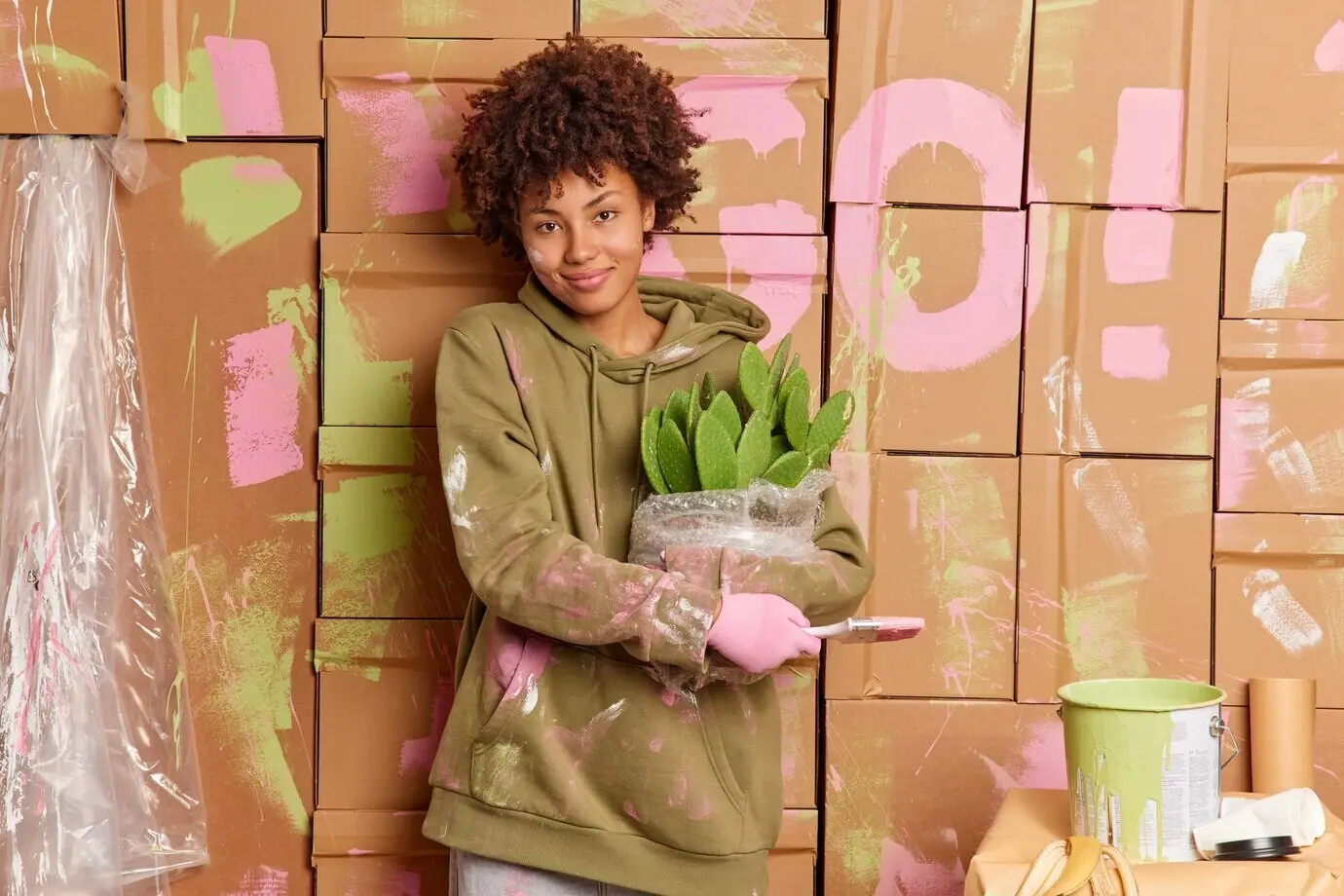 Satisfied female homeowner in a casual sweatshirt holds a paint brush and a potted cactus, busy with repairs in a new house, refurbishing the walls and standing with a glad expression. Interior renovation.